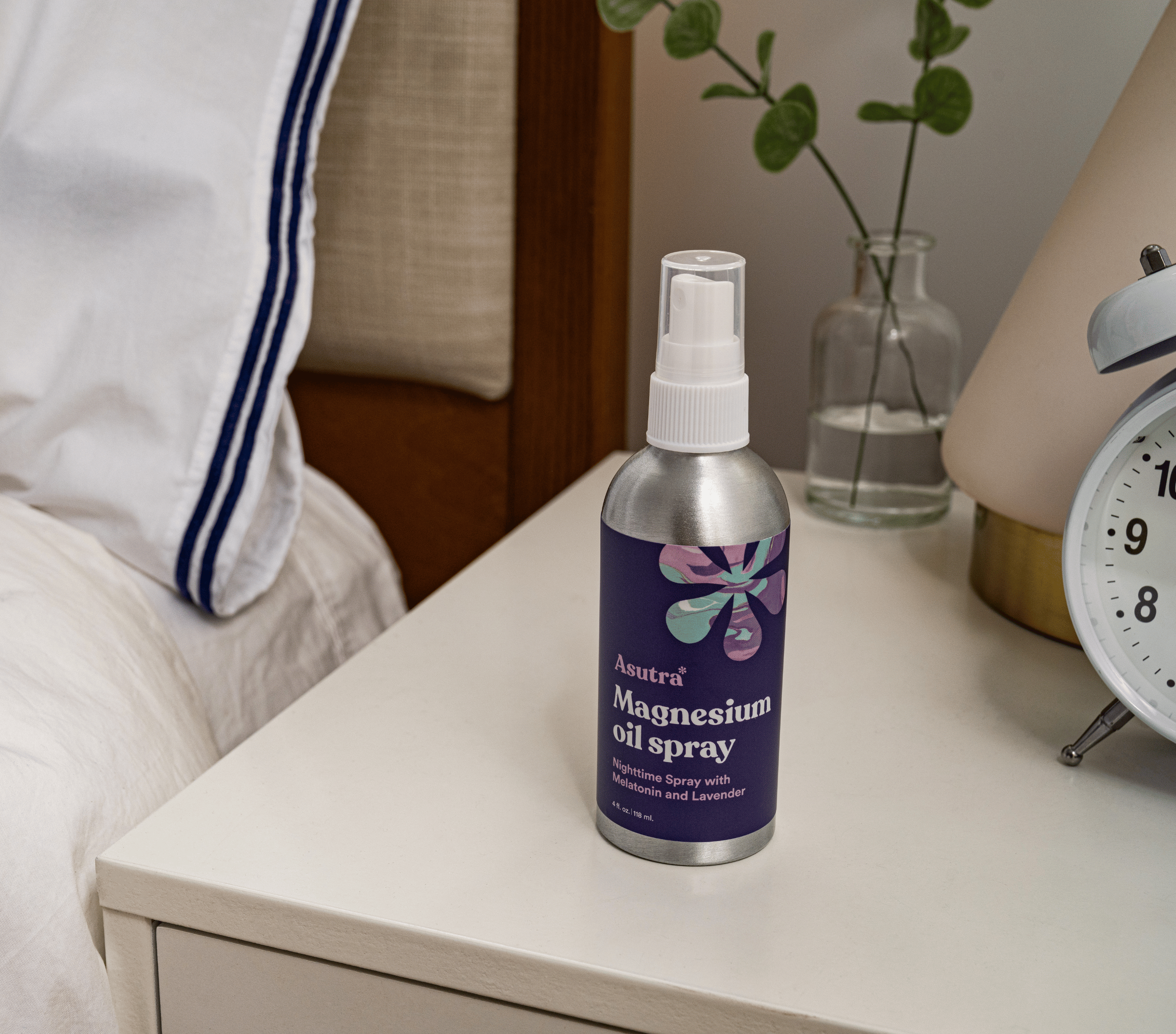 A silver spray bottle labeled "Magnesium Oil Spray with Melatonin and Lavender" on a white bedside table next to a bed.