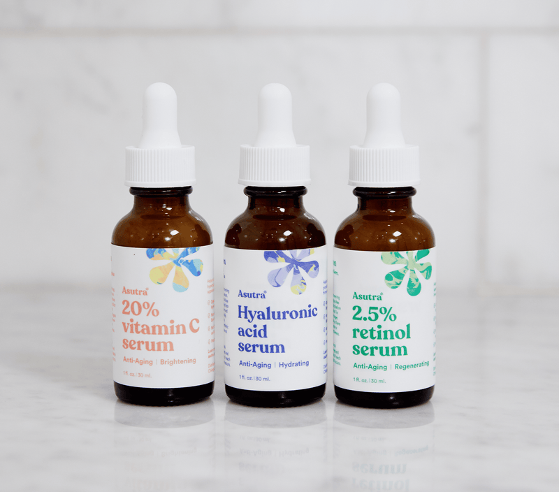 Three bottles of Asutra serums: 2.5% retinol serum, 20% vitamin C serum, and hyaluronic acid serum in white marble bathroom.