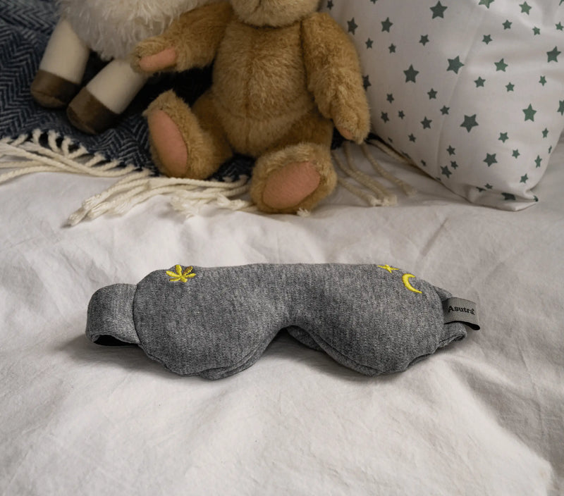 Gray kids lavender sleep mask with yellow embroidered designs, resting on a white bed with stuffed animals and pillows.