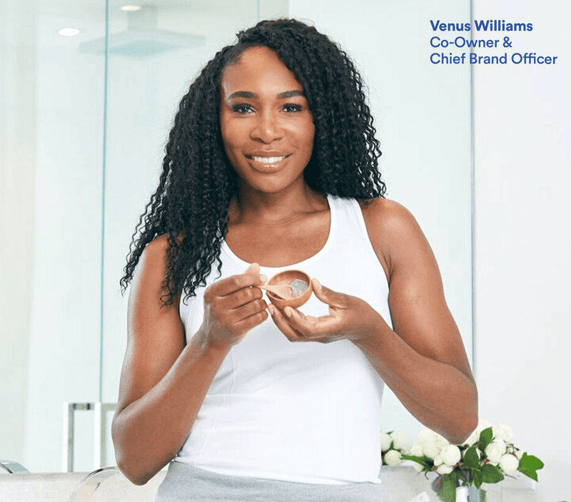 A woman holding a small wooden bowl of bentonite clay, smiling, in a bright, modern space with white flowers nearby.