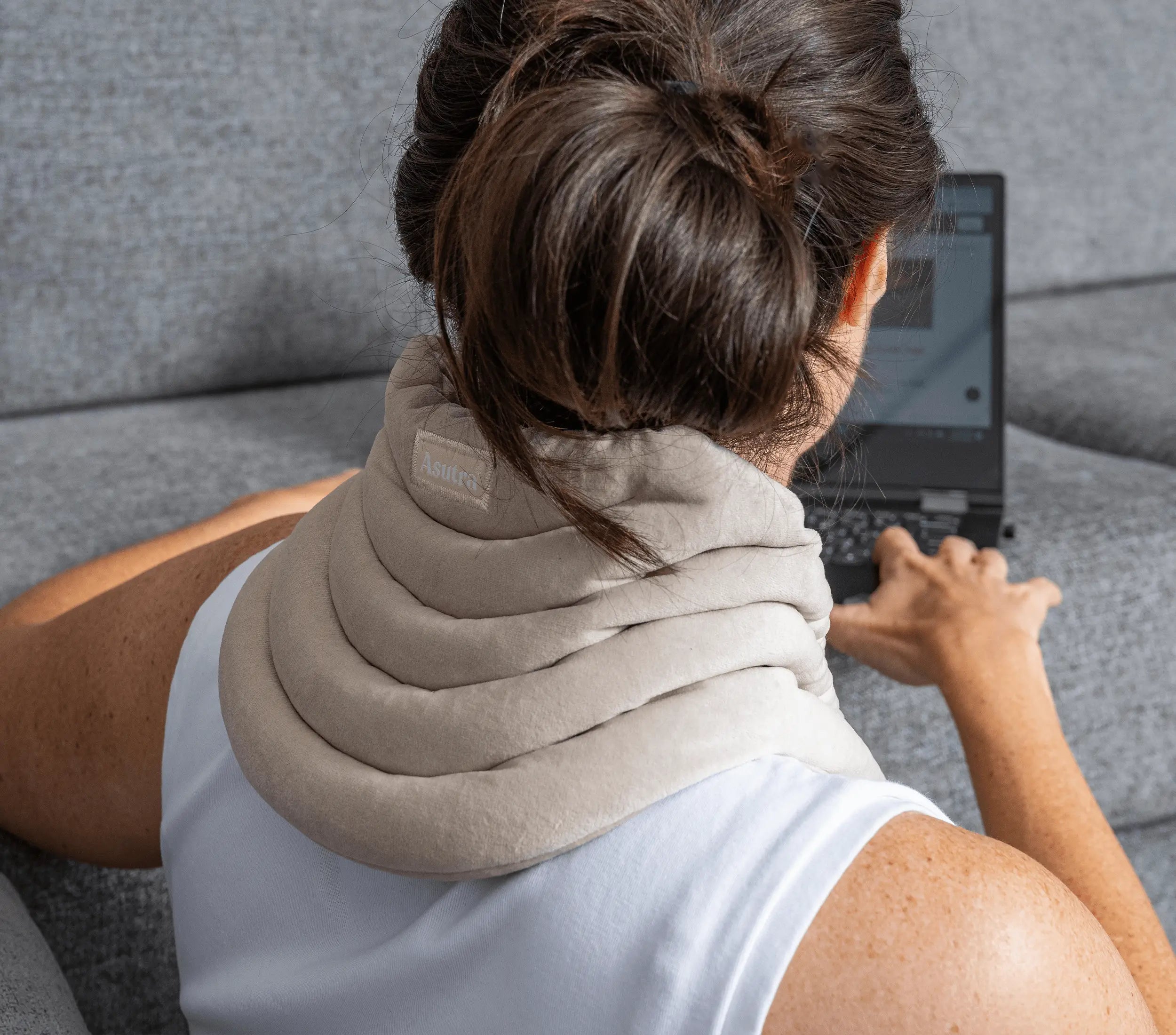 Cozy champagne-colored weighted neck wrap draped around a person's neck while they use a laptop on a gray sofa.