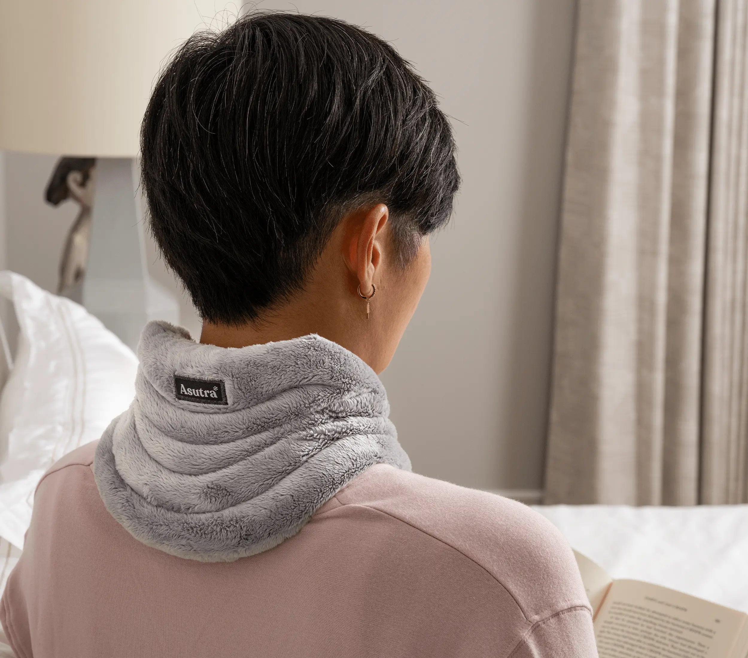 A woman wearing a cozy, plush gray weighted neck wrap while sitting on a bed, reading a book in a softly lit room.