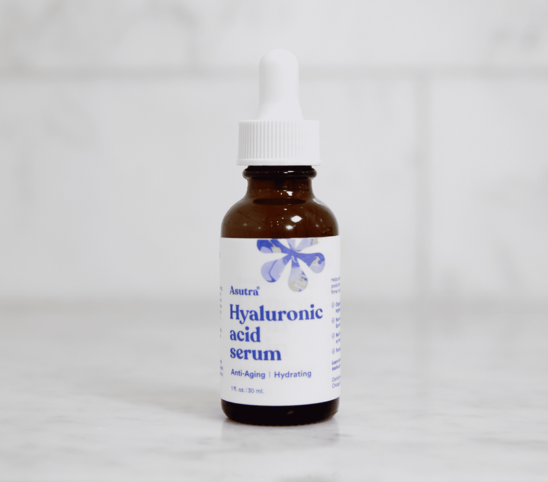 Amber glass dropper bottle of Asutra Hyaluronic Acid Serum with white label, blue text, and a floral design, on a white surface.
