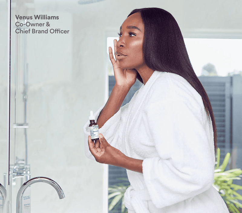 A woman in a white robe applying hyaluronic acid anti-aging serum, holding the bottle near her face in a bright, modern bathroom.