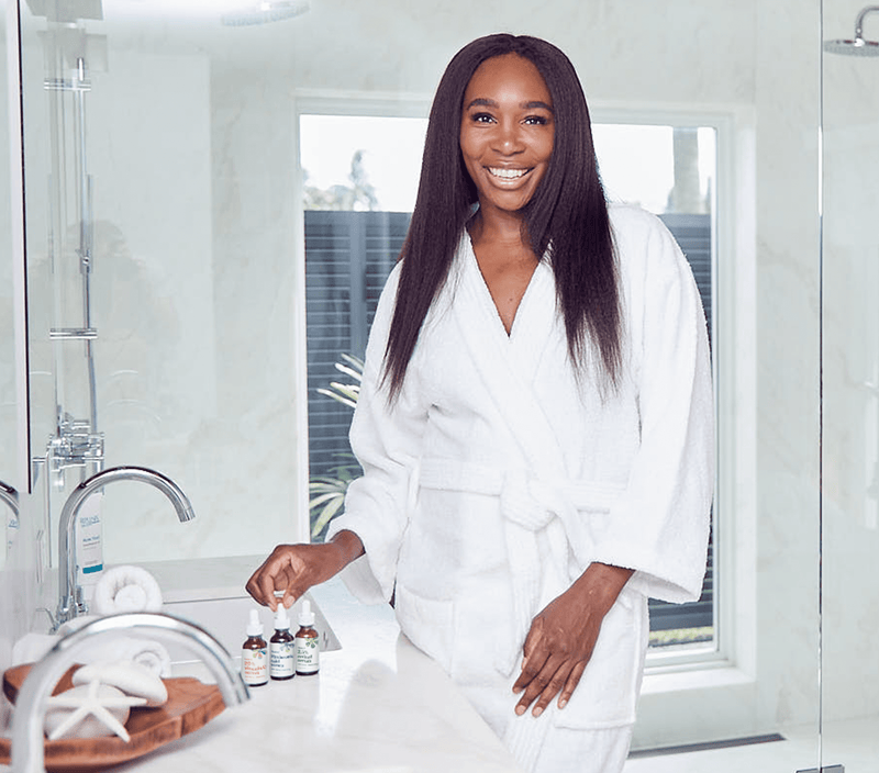 A woman in a white bathrobe in a bright bathroom, holding skincare serum bottles, promoting hyaluronic acid anti-aging serum.