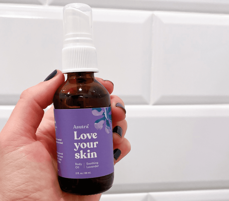 A hand holding a brown glass spray bottle of lavender-scented body oil with a purple label reading "Love your skin" against a white tiled background.