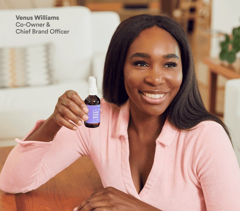 A smiling woman in a pink shirt holds a small brown bottle of lavender body oil with a spray top, showcasing the product.