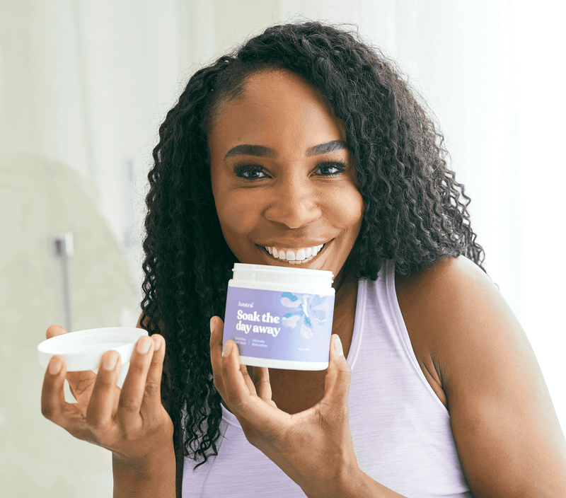 Smiling woman holding lavender relaxation bath salts container and scoop, promoting relaxation and self-care.