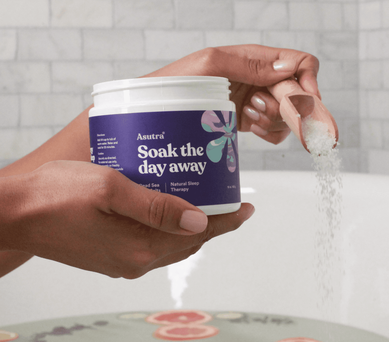 Hand holding a jar of Asutra Natural Sleep Therapy bath salts, pouring salts into a bowl for a relaxing bath.