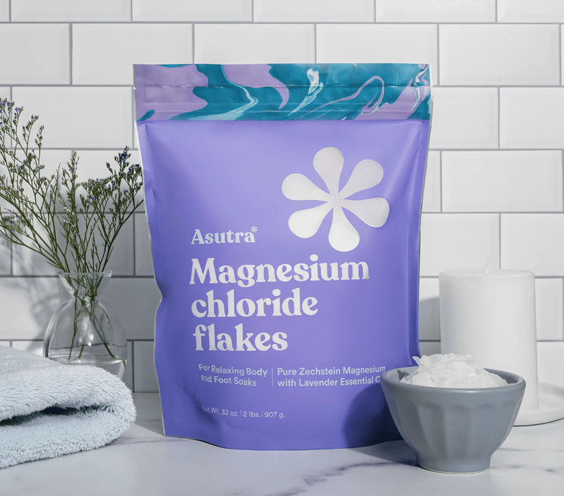 Lavender magnesium flakes in a purple resealable bag, displayed with a bowl of flakes, a towel, and a vase with flowers on a white marble surface.