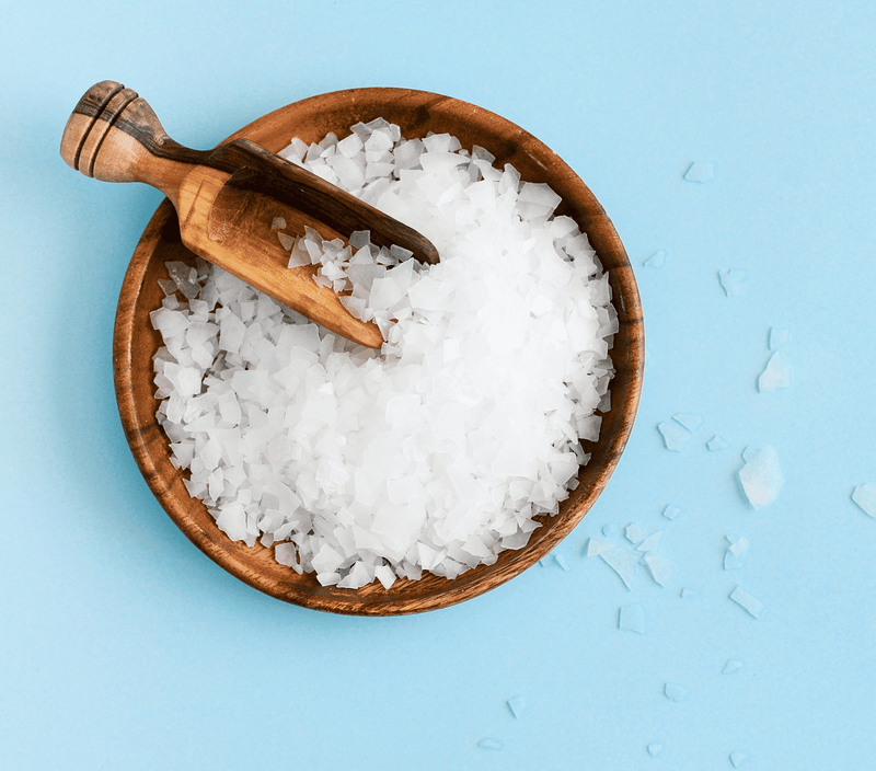 Natural magnesium chloride flakes in a wooden bowl with a scoop, set against a light blue background.