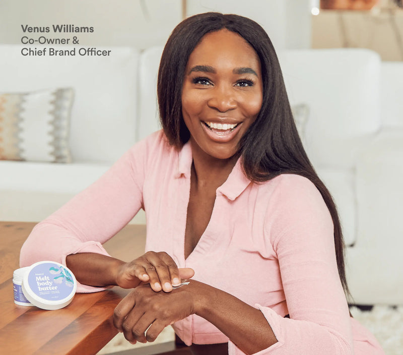 A woman applying lavender body butter with magnesium, smiling warmly, seated at a wooden table in a cozy, bright living room.