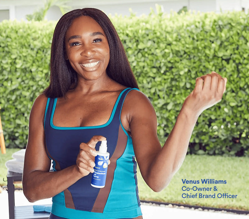 A woman smiling outdoors, holding a magnesium oil spray bottle in one hand and flexing her arm, with a green hedge background.