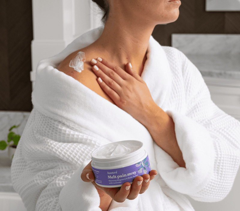 A person in a white robe applies Asutra's Melt Pain Away Lavender Body Butter on their shoulder, showcasing relaxation and self-care.