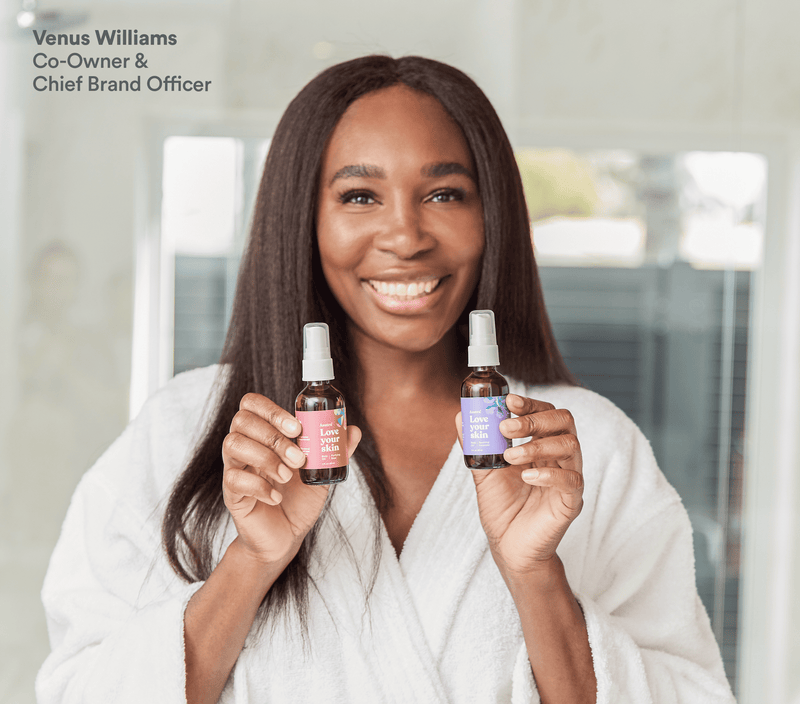 Smiling woman in a white robe holding rose and lavender body oil bottles, promoting a soothing skincare bundle.