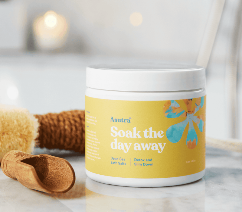 Container of Asutra Detox & Slim Down Bath Salts with a wooden scoop and textured towels on a marble surface.