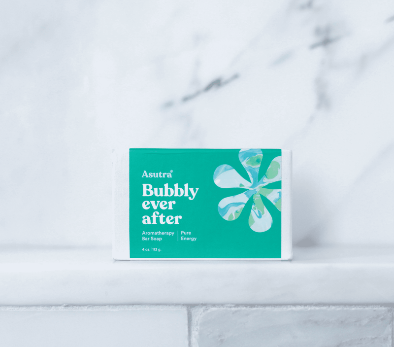 Bar soap with a teal and white packaging labeled "Asutra Bubbly ever after," featuring a floral design, on a white tiled surface.