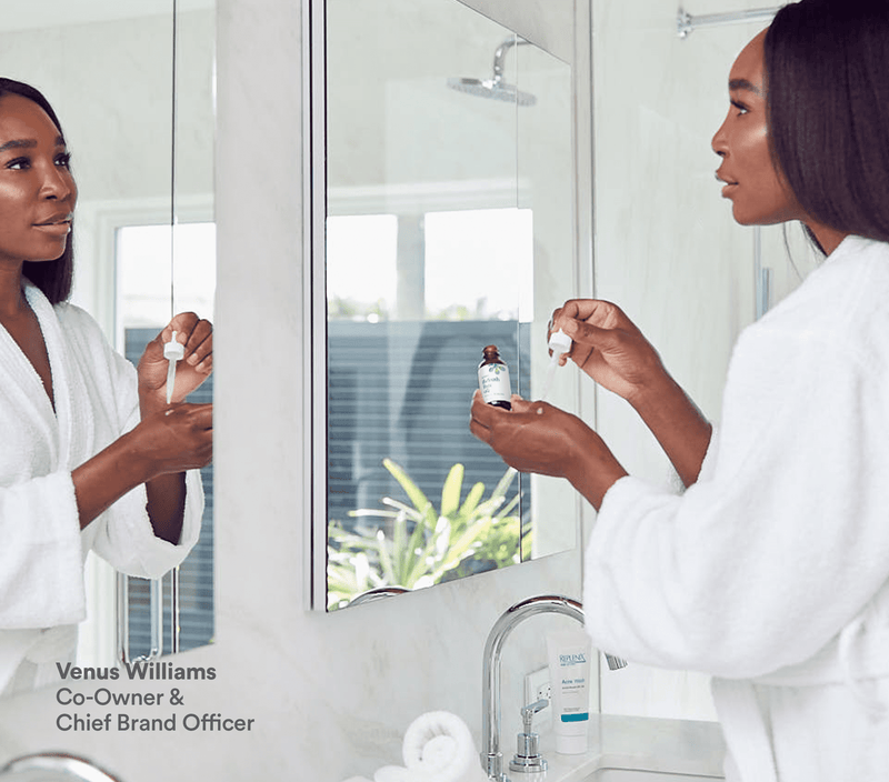 A woman in a white bathrobe applies Refresh Face Oil from a dropper in front of a mirror in a bright bathroom.