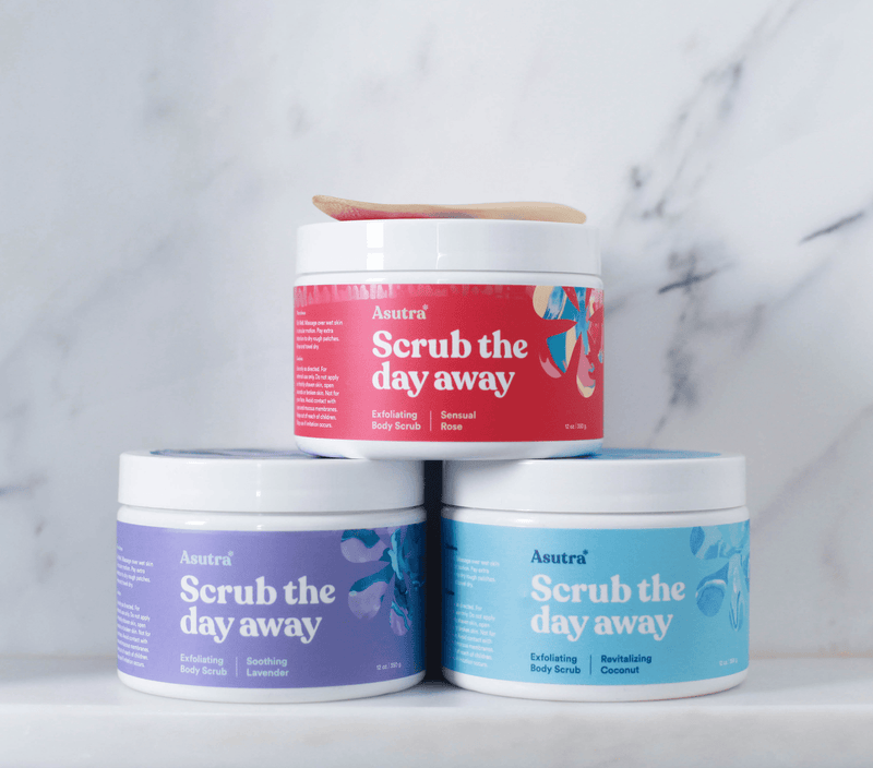 Three jars of Asutra exfoliating body scrubs in pink, lavender, and blue packaging, labeled "Scrub the day away" with scents: Sensual Rose, Soothing Lavender, and Revitalizing Coconut.