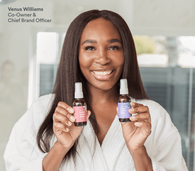 Smiling woman in a white robe holding two bottles of rose-scented body oil, promoting skincare and wellness products.