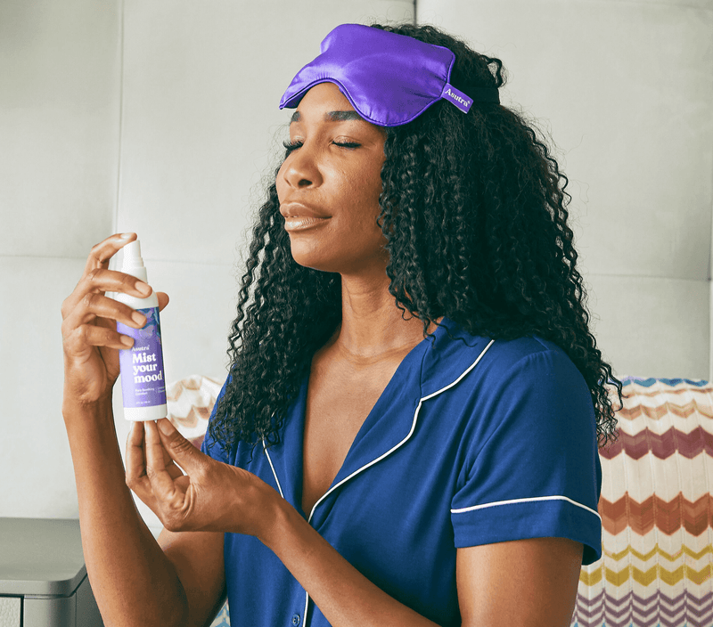 Woman with curly hair wearing purple sleep mask and blue pajamas, holding mist spray labeled "Mist your mood" for relaxation.