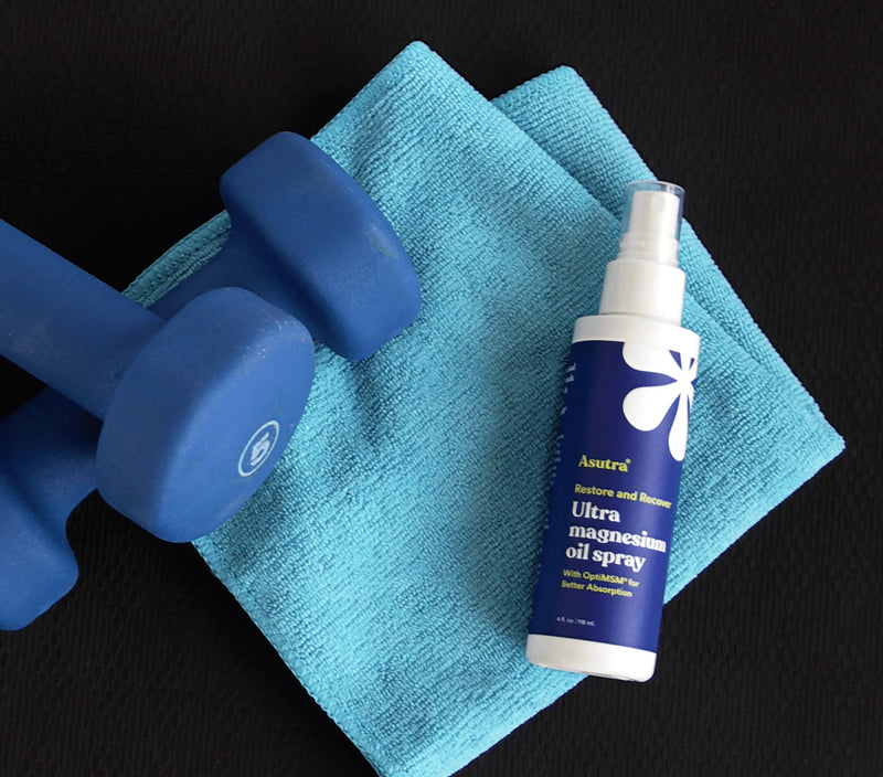 Blue dumbbells, a blue towel, and a white spray bottle of Ultra Magnesium Oil Spray on a black background.