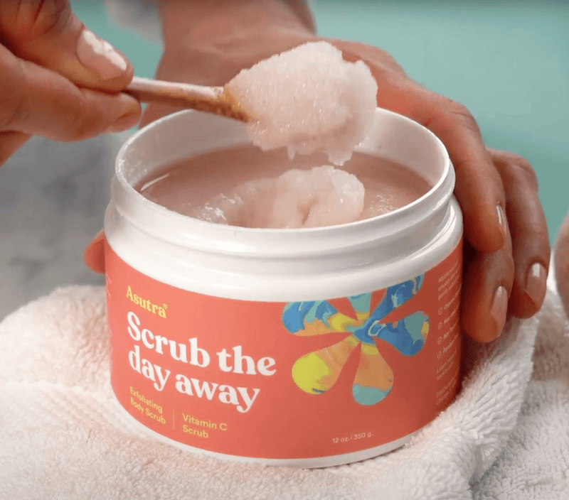 A person scooping pink Vitamin C body scrub from a jar labeled "Scrub the day away" with a wooden spatula, resting on a white towel.