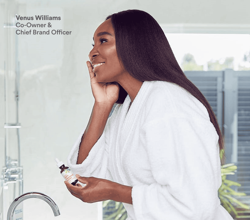 A woman in a white robe holding a Vitamin C anti-aging serum bottle, smiling in a bright, modern bathroom setting.