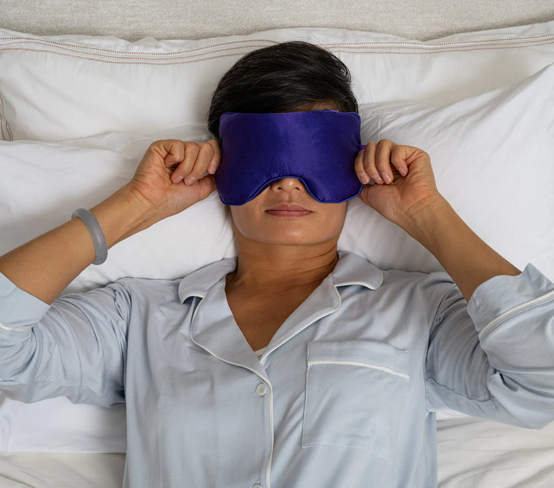 Woman lying in bed wearing a purple silk eye pillow, holding it over her eyes, with a purple gift box nearby.