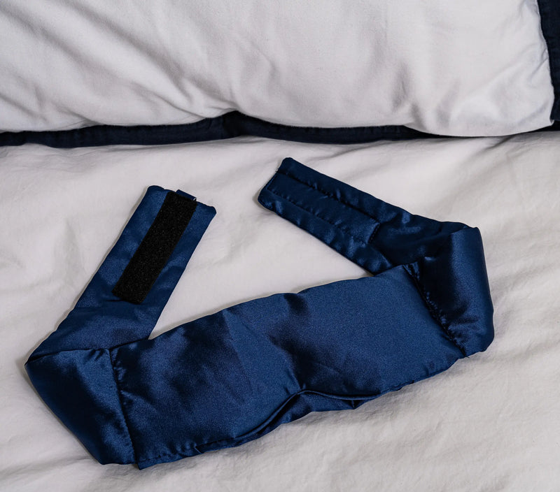 Navy sleep mask wrap with adjustable strap, made of silky fabric, resting on a white bedsheet.