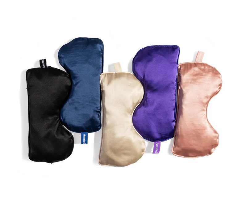 Set of five silk eye pillows in black, navy blue, cream, purple, and rose gold, each in a purple gift box.