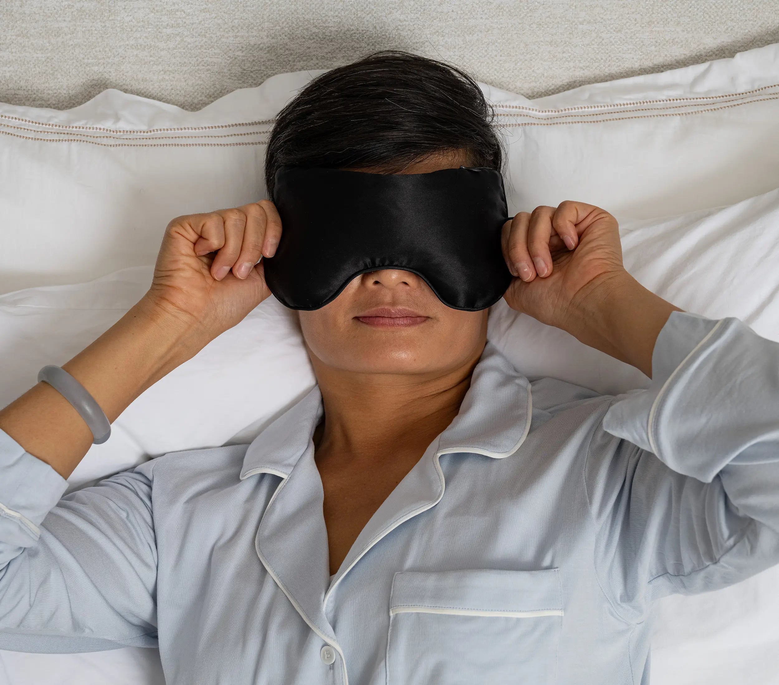 Woman lying in bed wearing a black silk eye pillow, relaxing with eyes covered for rest or meditation.