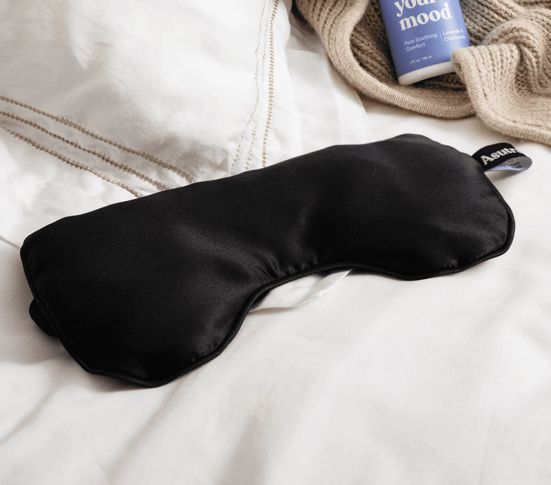 Black silk eye pillow resting on white bedding, with a beige sweater and lavender-scented sleep spray nearby.