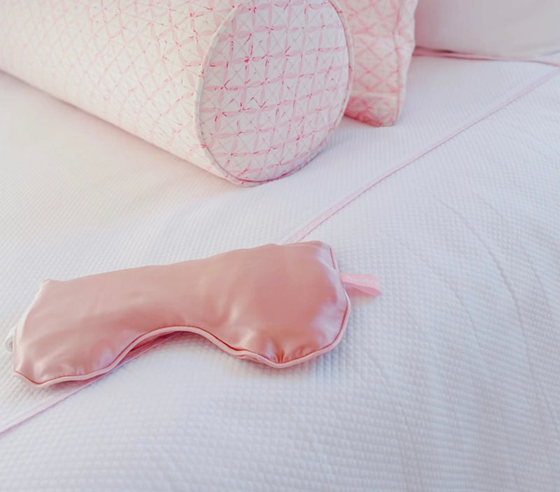 Pink silk eye pillow with a matching pink satin sleep mask resting on a white textured bedspread.