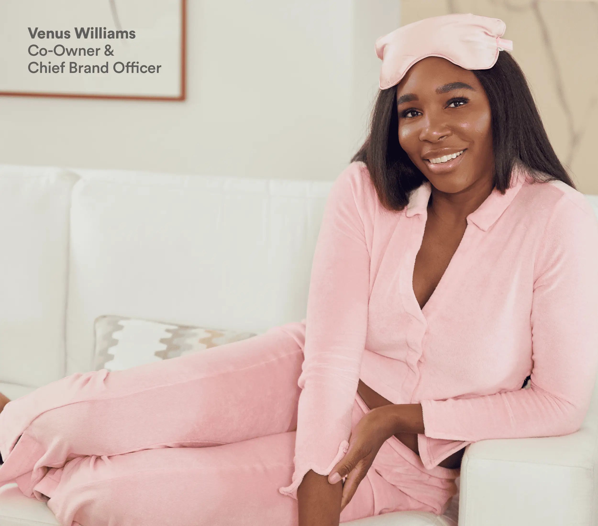 Pink silk eye pillow resting on a woman's forehead, she is sitting on a white couch wearing matching pink loungewear.