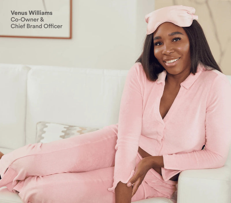 Pink silk eye pillow resting on a woman's forehead, she is sitting on a white couch wearing matching pink loungewear.