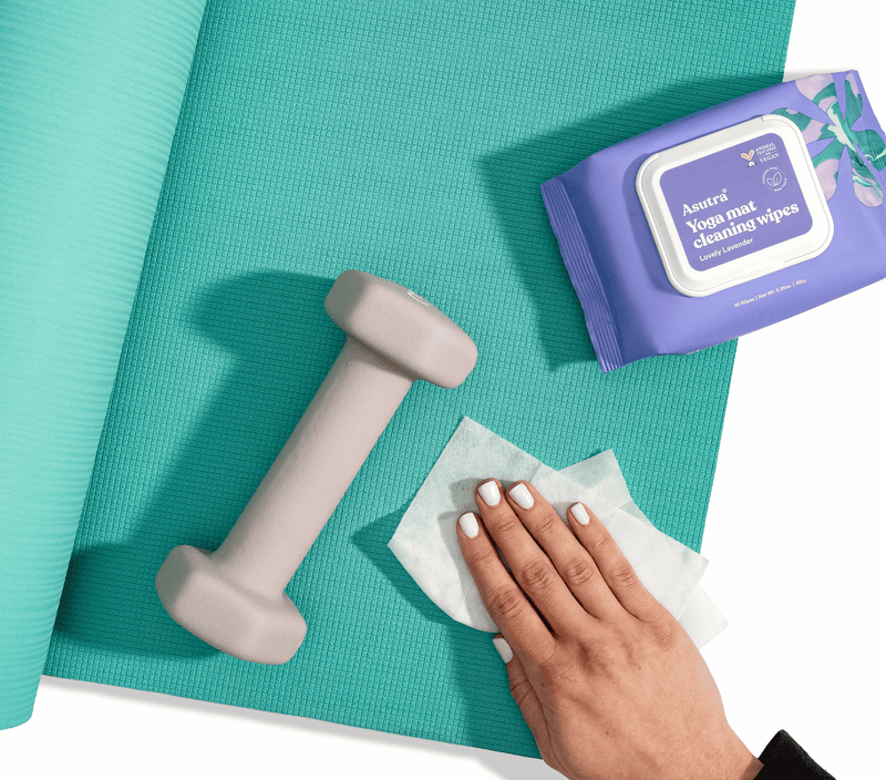 Lavender yoga mat cleaning wipes pouch, a hand wiping a yoga mat, a small dumbbell, and a teal yoga mat on a white background.
