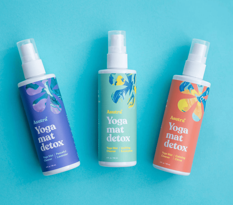 Three bottles of Asutra yoga mat cleaner spray with colorful labels, designed for cleaning and detoxing yoga mats.