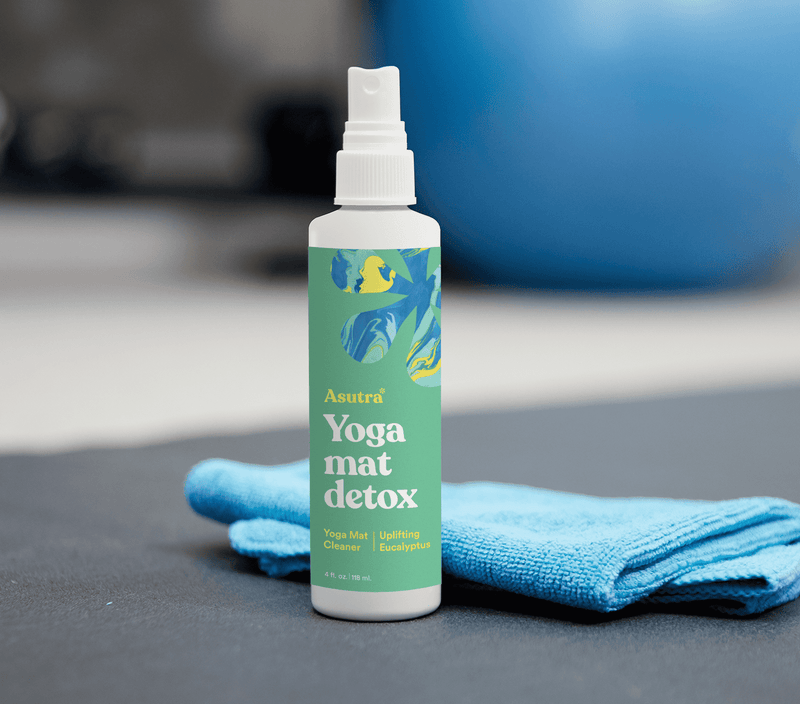 A white spray bottle of Uplifting Eucalyptus Yoga Mat Cleaner with a blue microfiber cloth on a gray surface, blurred background.