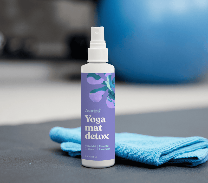 A white spray bottle labeled "Asutra Yoga Mat Detox" with peaceful lavender design, placed on a blue towel on a yoga mat.