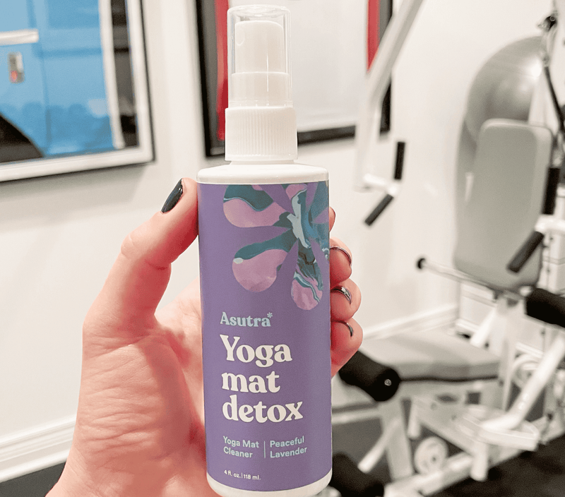 Bottle of Asutra Peaceful Lavender Yoga Mat Cleaner with a spray nozzle, held in front of gym equipment.