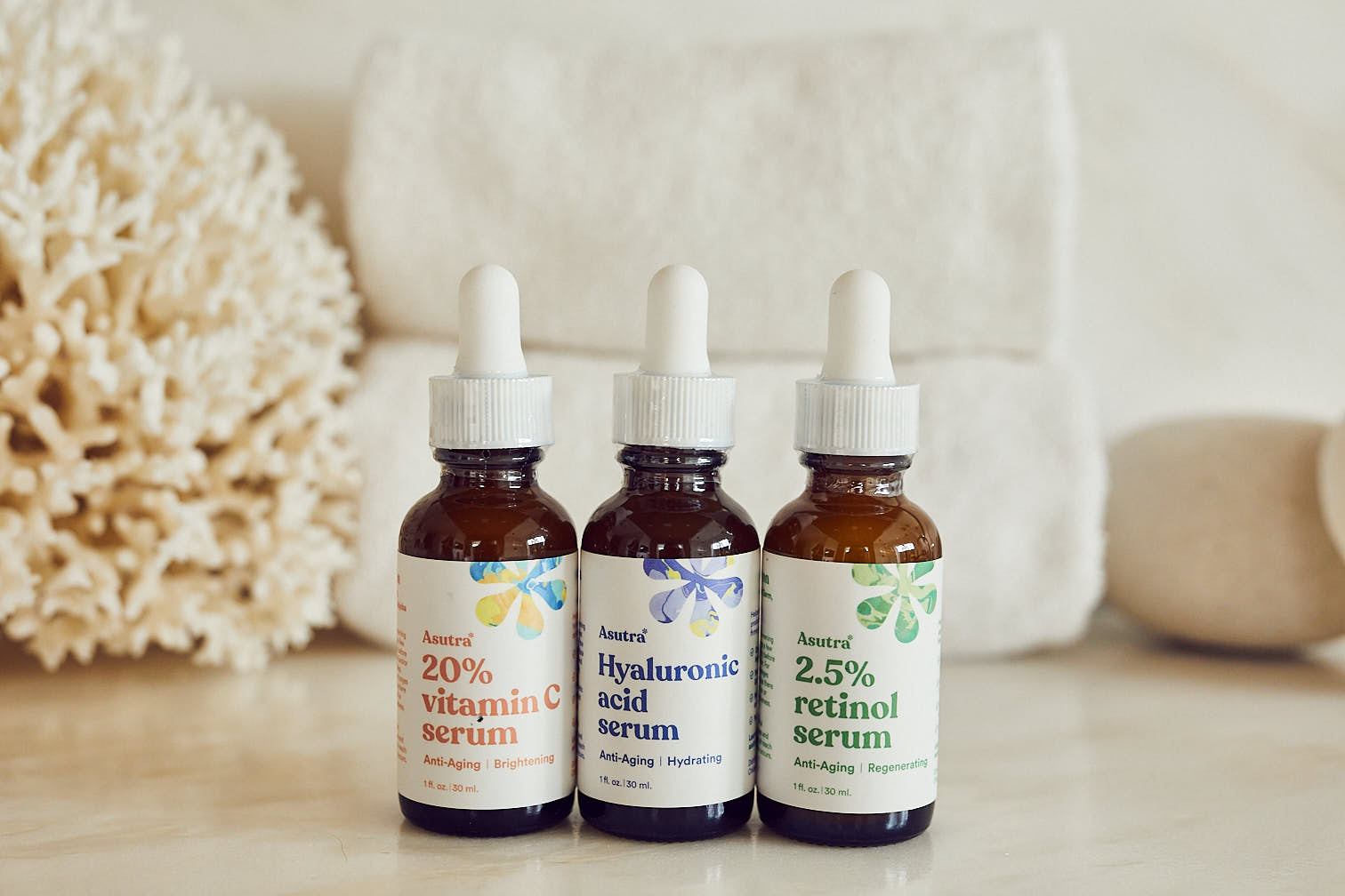Three bottles of Asutra serums: 2.5% retinol serum, 20% vitamin C serum, and hyaluronic acid serum on bathroom counter