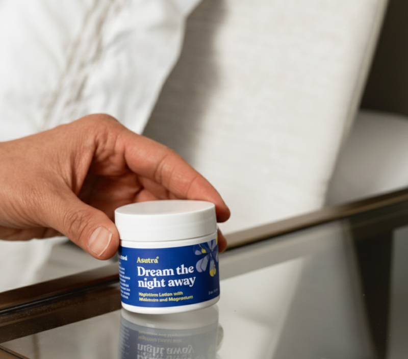 A person holds a jar of Asutra's Dream the Night Away Lotion on a table, emphasizing its calming benefits for sleep.