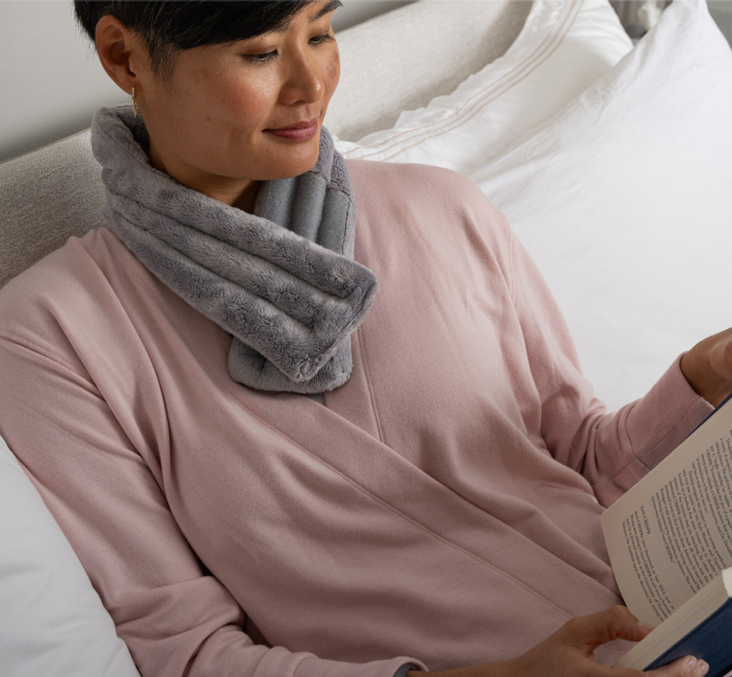 A woman relaxes in bed wearing a plush, cozy gray weighted neck wrap, while reading a book.