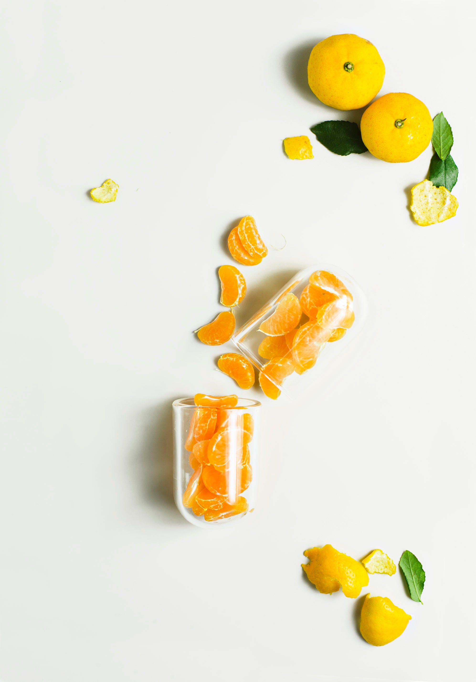 Fresh peeled and whole oranges scattered with glass cups, representing vitamin C, against white background.