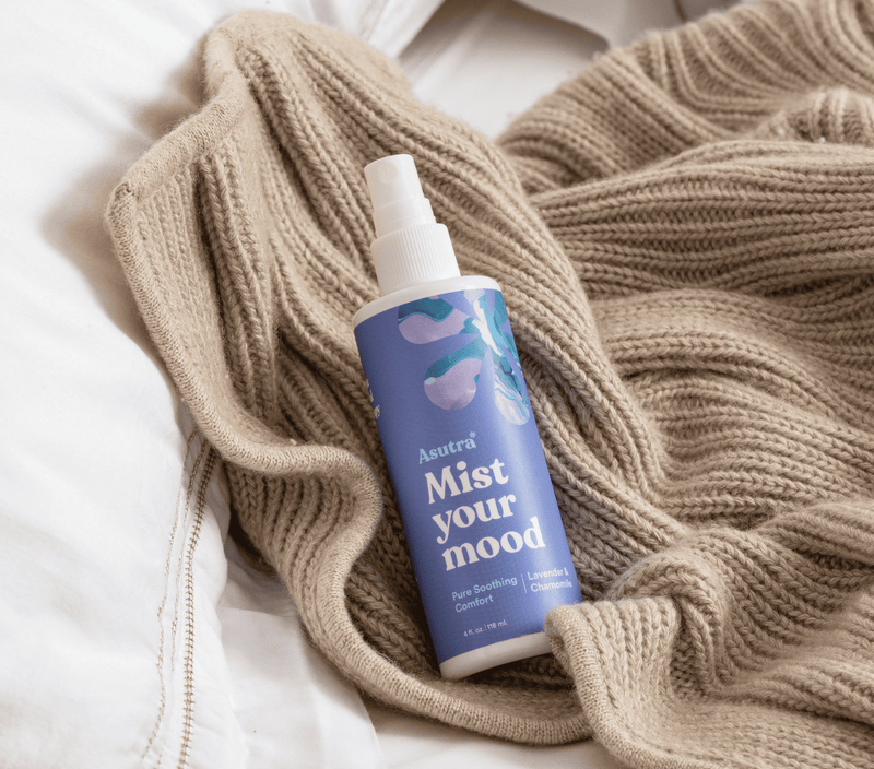 A bottle of Asutra Mist Your Mood lavender aromatherapy spray rests on a cozy beige knit blanket. 