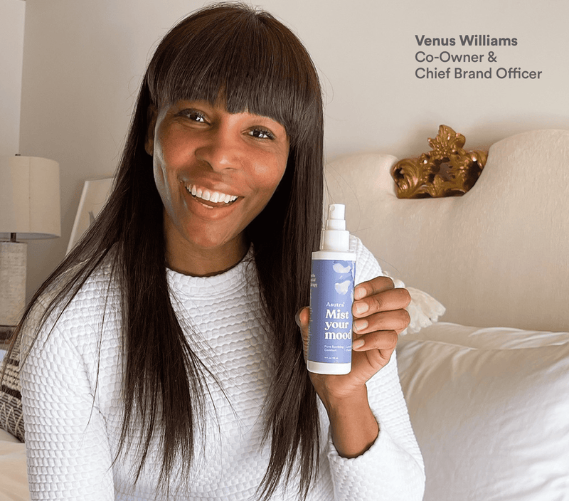 Venus Williams smiling sitting on a bed and holding Asutra Mist Your Mood lavender aromatherapy mist. 