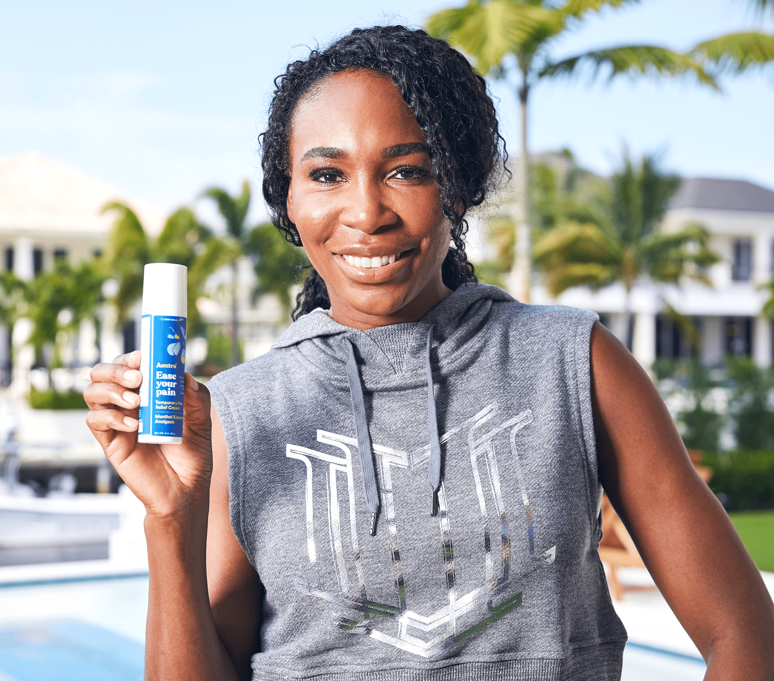 Venus Williams smiles while holding a bottle of Asutra Ease Your Pain cream, standing outside with palm trees in the background.